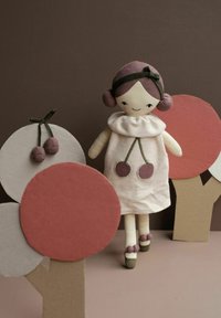 Weiche Stoffpuppe mit pinkfarbenem Haar, trägt ein weißes Kleid mit Kirschapplikationen. Im Hintergrund sind Pappbäume mit rundem Laub zu sehen.