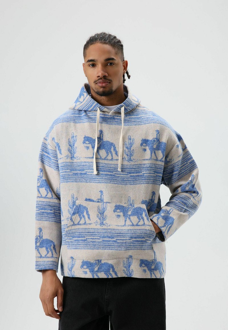 Capuchontrui met een beige basis, blauw paarden- en cactuspatroon, een voorzak en een sluiting met trekkoord. Knusse textuur.