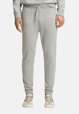 Man draagt lichtgrijze joggingbroek met trekkoord in de taille en omgeslagen enkels, gecombineerd met beige en witte lage sneakers.