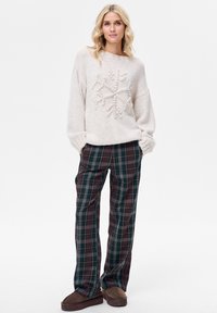 Jasnobeżowy sweter z fakturą przypominającą płatek śniegu, zestawiony z ciemnymi, kratkowymi spodniami w zielone, bordowe i żółte akcenty. Brązowe wsuwane buty.