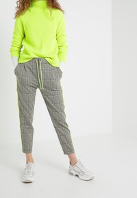 Jasnozielony neonowy prążkowany sweter z golfem w połączeniu z wzorzystymi szarymi zwężanymi spodniami z neonowymi paskami po bokach oraz białymi trampkami.