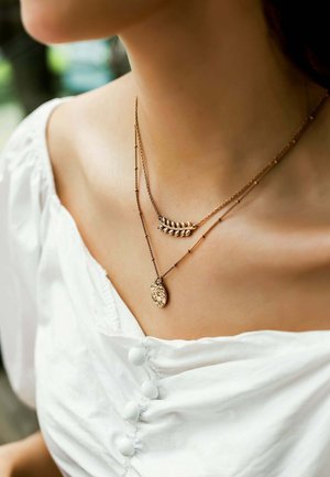 Deux colliers en or superposés sur le cou d'une femme, l'un avec un pendentif en forme de feuille et l'autre avec un pendentif rond martelé, portant un chemisier blanc.