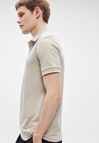 Μπεζ πόλο μπλουζάκι με κοντά μανίκια από υφασμάτινη υφή, με κλασικό γιακά και αντίθετες λεπτομέρειες στα μανίκια. Χωρίς εμφανή λογότυπα.