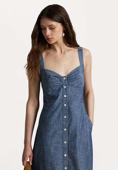 Robe en denim bleu avec un devant à boutons, un buste froncé, des bretelles larges et des poches, présentant une texture lisse et une silhouette ajustée.