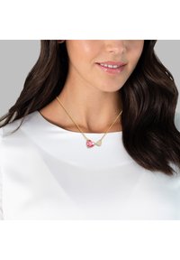 Guldkedja med två hänganden: ett rosa hjärta och ett mindre hjärta med en texturerad mönster. Bärs över en vit blus.