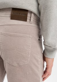 Close-up van de beige corduroy broekzak aan de achterkant met een bruine leren label "Andrew James" gedragen met een lichtgrijze sweatshirt.