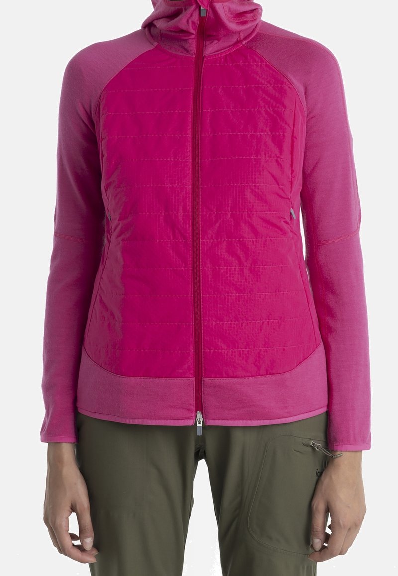 Femme portant une veste rose zippée avec des panneaux avant matelassés et des manches unies, associée à un pantalon vert olive avec une poche zippée.