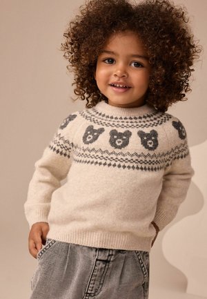 Lächelndes Kind mit lockigem Haar, das einen cremefarbenen Pullover mit grauen Bärengesichtern und gemusterten Designs trägt, kombiniert mit grauer Hose.