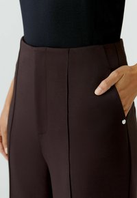 Dunkelbraune Hose mit glatter Textur, ausgestattet mit einer Seitentasche und einem kleinen Metall-Logo-Detail nahe der Tasche. Hohe Taille.