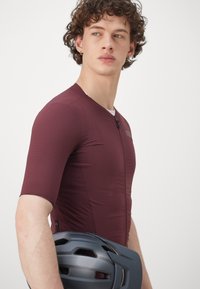 Burgunderfarbene Radtrikot mit kurzen Ärmeln, strukturiertem Stoff, tailliertem Schnitt und einem Reißverschluss; wird mit einem grauen Helm gehalten.