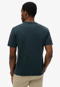 Man die met zijn rug naar de camera staat, gekleed in een donkerblauw T-shirt met korte mouwen en beige broek, met een horloge om zijn linkerpols, staand tegen een witte achtergrond.