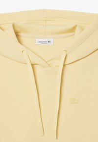 Gelbe Lacoste-Hoodie mit lockerem Schnitt, verstellbarem Kordelzug an der Kapuze und gesticktem Logo auf der Brust sowie einer glatten Stofftextur.
