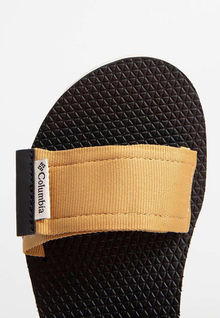 Semelle de sandale noire texturée avec une large sangle en tissu beige présentant une étiquette du logo Columbia sur le côté.