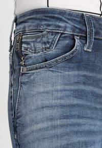 Acercamiento a unos pantalones de mezclilla azul con un pequeño bolsillo con cremallera y bucles para el cinturón visibles, usados con una blusa blanca metida por dentro.