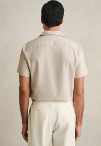 Camisa beige de manga corta con cuello clásico, de tela suave y detalle en el yugo trasero, combinada con pantalones ajustados de un color claro.