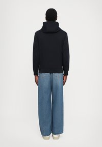 Sudadera con capucha de color navy y corte relajado, combinada con jeans de pierna ancha y zapatillas blancas, vista desde atrás.