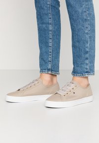 Beige Sneaker mit niedrigem Schaft, weißen Sohlen und gestreiften Schnürsenkeln, getragen zu blauen Jeans, zeigen Seiten- und Vorderansichten beider Schuhe.