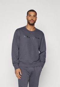Grauer Pullover mit rundem Hals und langen Ärmeln, ausgestattet mit einem gestickten "Champion"-Logo in kontrastierender Farbe über der Brust. Weicher Stoff.