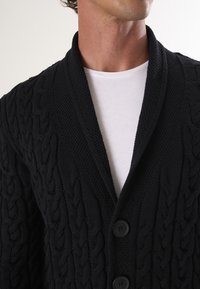 Zwarte gebreide cardigan met een textuur kabelpatroon, sjaalkraag en twee knopen. Draag het over een wit shirt.
