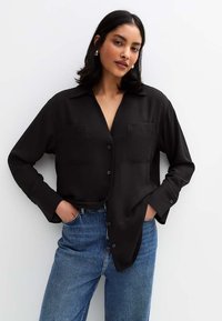 Camisa negra de manga larga y botones con una textura suave, que presenta dos bolsillos frontales y un corte relajado, combinada con unos jeans azules de cintura alta.