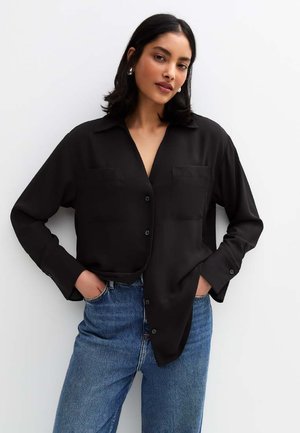 Chemise noire à manches longues et à boutons, avec une texture douce, présentant deux poches avant et une coupe décontractée, associée à un jean bleu taille haute.