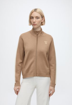 Femme aux cheveux blonds courts portant une veste zippée marron clair avec des rayures blanches sur les manches et un pantalon blanc, debout devant un fond uni.