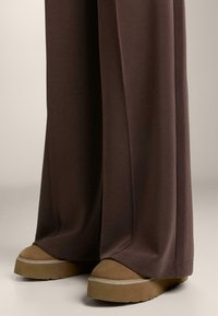 Široke vijolično-rjave hlače se raztezajo čez svetlo rjave platformne čevlje z teksturirano površino in večplastno zasnovo podplata.