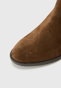 Bottine en suede marron avec un bout rond, texture lisse, et une semelle en caoutchouc noire comprenant un détail cousu le long du bord.