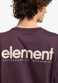 Camiseta de algodón morada con un gran logo blanco en la parte posterior que dice "element SKATEBOARD CO. WORLDWIDE". Diseño de manga corta y cuello tipo crew.