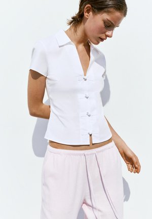 Ženska nosi prilegajočo belo kratko srajco z V-izrezom in dekorativnimi vozli, kombinirano s svetlo rožnatimi hlačami z vrvico.
