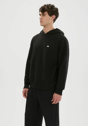 Joven con sudadera negra con capucha y pantalones negros a juego, de pie frente a un fondo blanco liso.