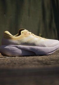 Ανοιχτό μπεζ και λευκό παπούτσι τρεξίματος Adidas με λεπτομέρεια σόλας "Dreamstrike+", τοποθετημένο σε τραχύ έδαφος με σκοτεινό θολό φόντο.
