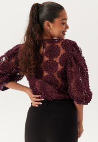 Genomskinlig lila blus med strukturerade blommönster. Löst sittande trekvartsärmar och detalj med nyckelhål i ryggen. Bärs över svarta byxor.