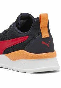 Chaussure de sport noire avec une bande latérale rouge, un accent orange au niveau du talon, un dessus en tissu texturé et une semelle intermédiaire blanche avec un motif surélevé.