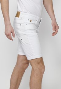 Pantalones cortos de mezclilla blanca con dobladillo enrollado, que presentan un parche en la cintura trasera, herrajes metálicos y un diseño de ajuste slim.