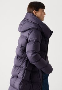 Lange paarse pufferjas met hoge kraag, gewatteerd ontwerp en rits-sluiting. Voorzien van een capuchon en zijzakken.