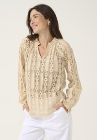 Lace blouse in beige featuring a V-neckline, long sleeves, textured patterns with circular cutouts, and a relaxed fit.