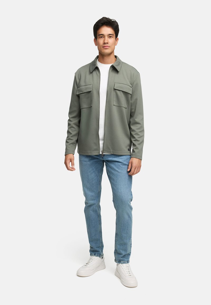 Vihreä fleece-takki, jossa kaksi etutaskua, päällä valkoinen paita, yhdistetty sinisiin farkkuihin ja valkoisiin lenkkareihin. Malli seisoo suorassa.