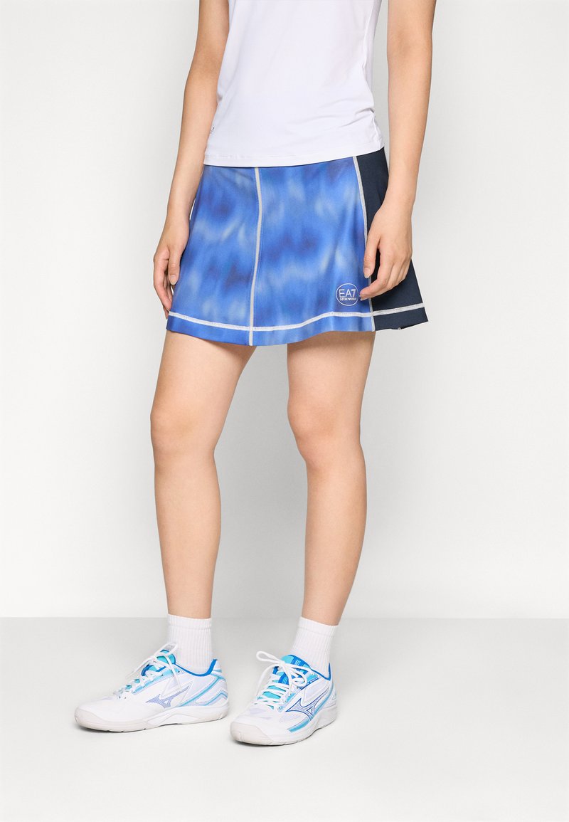 Saia atlética azul e marinha com um padrão tie-dye, apresentando costuras prateadas e um logo EA7. Usada com tênis esportivos brancos e azuis.