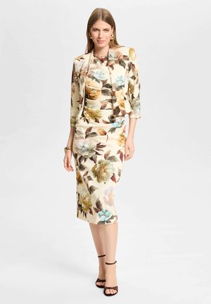Femme portant une robe fleurie couleur crème et un blazer assorti, accessoirisée de boucles d’oreilles dorées, un bracelet et des talons noirs à bride à la cheville.