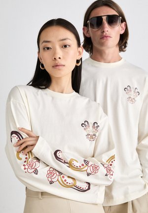 Cream long-sleeve t-shirts with floral and abstract designs in red and gold. Features a crew neck and patterned sleeves.