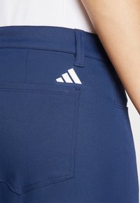 Temno modre hlače z zadnjim žepom, na katerem je bel Adidasov logotip. Tkanina je videti gladka, oblikovana s čistim in urejenim krojem.