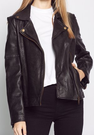 Femme portant une veste en cuir noir de motard par-dessus un t-shirt blanc, une main dans la poche et cheveux raides visibles.