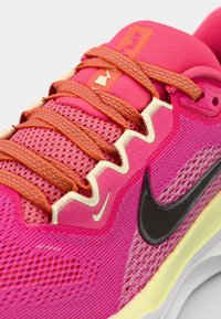 Pink athletic shoe with a mesh upper, contrasting orange laces, and a black logo. Features textured overlays and a cushioned sole.