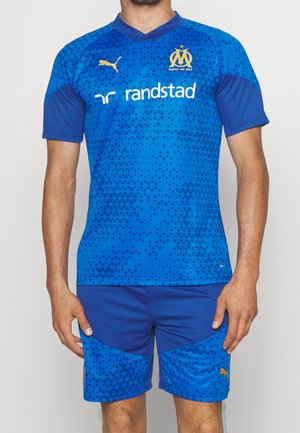 Mand iført blå Olympique de Marseille fodboldtrøje med trekantsmønster, gyldne Puma-logoer og Randstad sponsor på fronten.