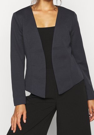 Woman wearing a fitted, open-front dark gray blazer over a black top and black pants, shown from neck to hips.