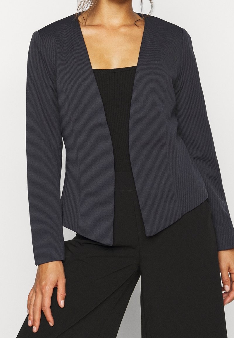 Femme portant un blazer cintré gris foncé ouvert sur un haut noir et un pantalon noir, visible du cou aux hanches.