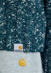 Teal gebreide trui met witte stippen, voorzien van een Carhartt-label en een geel geborduurd logo op een grijze onderhemd.