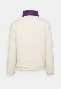 Kurtka z polaru w kolorze kremowym z fioletowym kołnierzem. Charakteryzuje się miękką teksturą i luźnym krojem, z długimi rękawami i prostym cięciem przy dolnym brzegu.