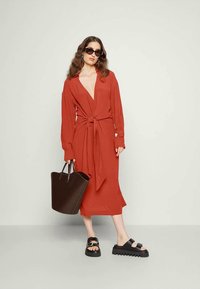 Mulher vestindo um vestido envolvente laranja-oxido com mangas longas, sandálias plataforma pretas, óculos de sol, brincos azuis, segurando uma grande mala preta.